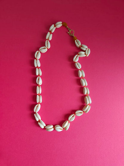 Long length cowrie shell and glass bead necklace on a pink background 