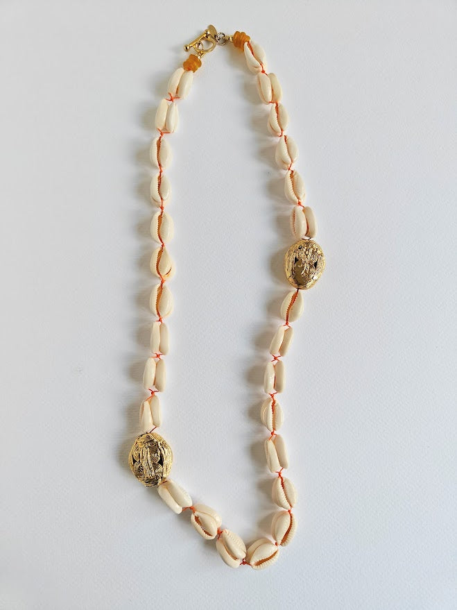 Long necklace with cowrie shell beads, large gold brass beads and accents of yellow recycle glass beads on a light gray background