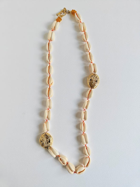Long necklace with cowrie shell beads, large gold brass beads and accents of yellow recycle glass beads on a light gray background