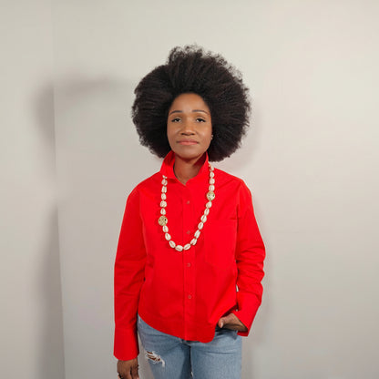 Person wearing a red shirt and a long cowrie shell necklace which has accents of large gold brass beads in it and  blue jeans against a light grey  background 