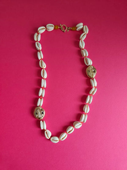 Double backed cowrie shell necklace with large brass beads, accents  of yellow recycled glass beads  on a pink background