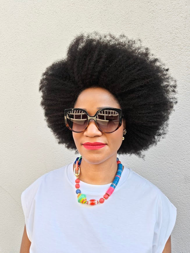 Person with large afro wearing sunglasses and a colorful necklace against a plain background