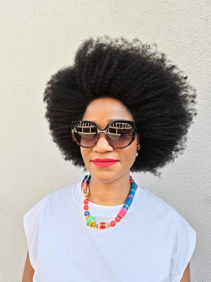 Person with large afro wearing sunglasses and a colorful necklace against a plain background