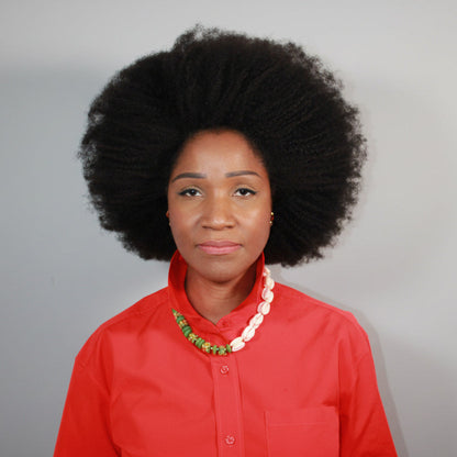 Woman wearing a red shirt with a  short length cowrie shell necklace, she's standing against a plain grey background