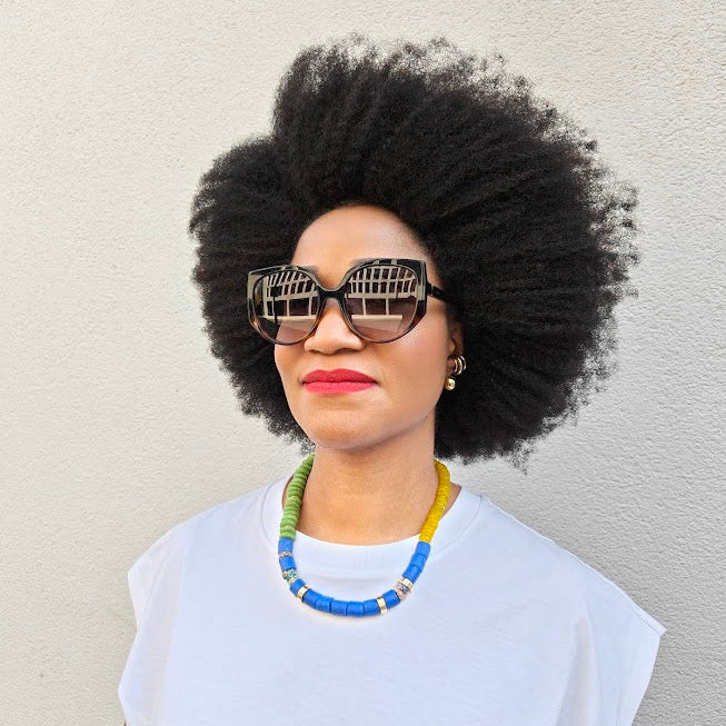 Person with an afro wearing sunglasses and a colorful necklace against a plain background