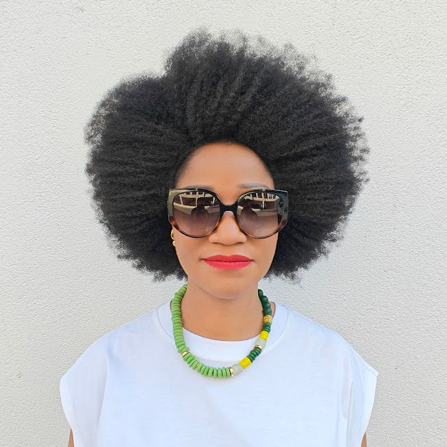 Woman with an afro wearing a beaded necklace, sunglasses and a white top against a light background