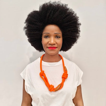 Woman with an afro wearing a white top and orange necklace with whisky colour leather trim against a light background