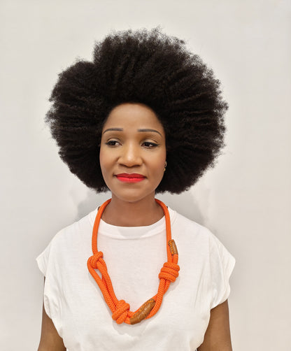 Woman with an afro wearing a white top and orange necklace with whisky colour leather trim against a light background