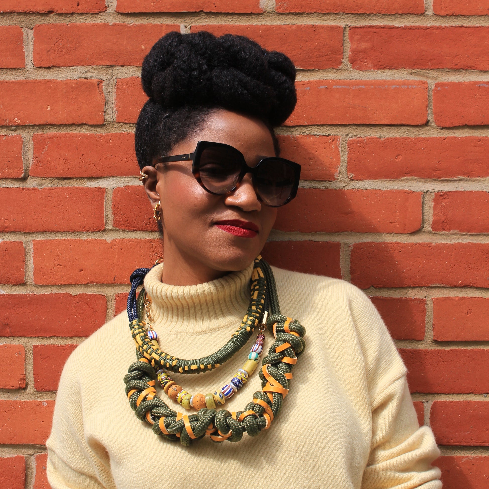 Woman wearing sunglasses and a beige sweater with colorful necklaces against a brick wall.