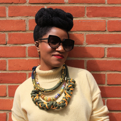 Woman wearing sunglasses and a beige sweater with colorful necklaces against a brick wall.