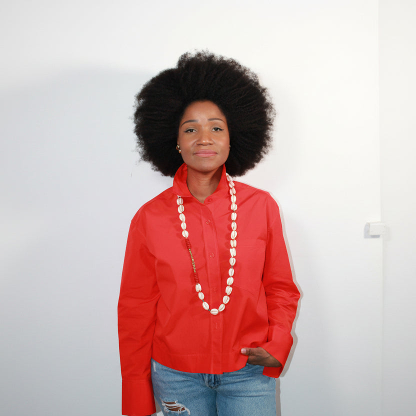 Person wearing a red shirt with a long cowrie shell necklace white buttons on a plain background