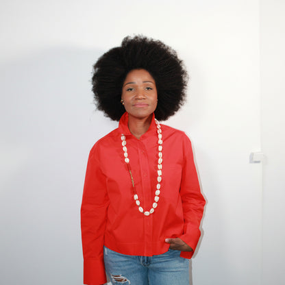 Person wearing a red shirt with a long cowrie shell necklace white buttons on a plain background